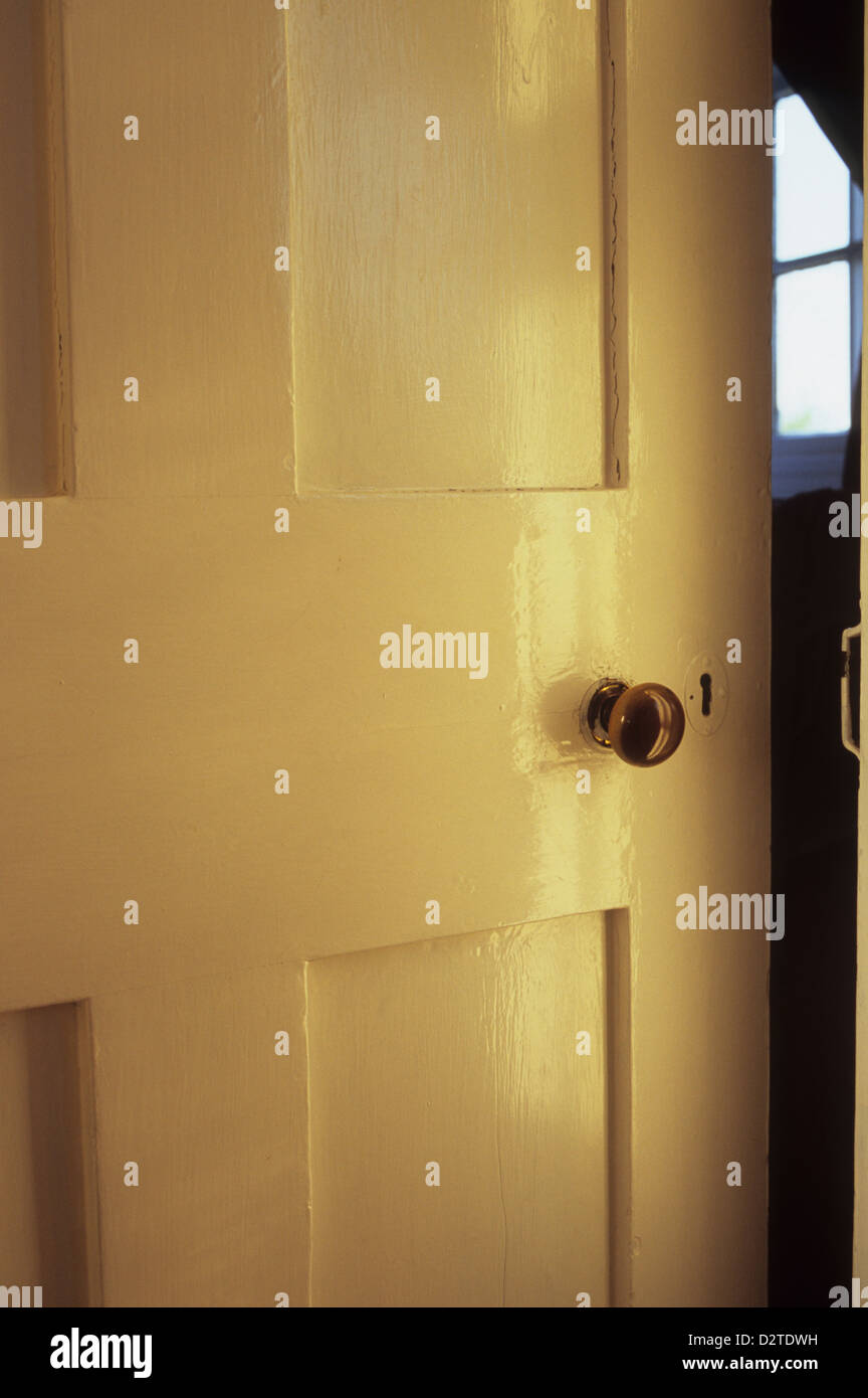 Dettaglio del vecchio interno con pannelli in legno porta dipinta di bianco con manopola in ottone in piedi socchiusa per rivelare la piccola finestra Foto Stock