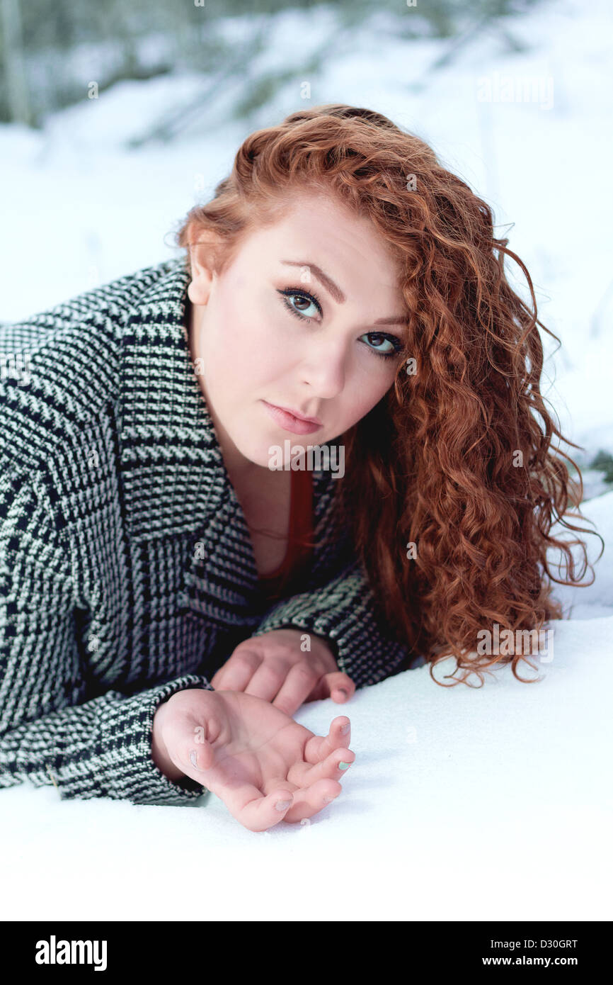 Bella giovane donna con ricci capelli rosso e bianco e nero cappotto a spina di pesce si trova su stomaco nella neve Foto Stock