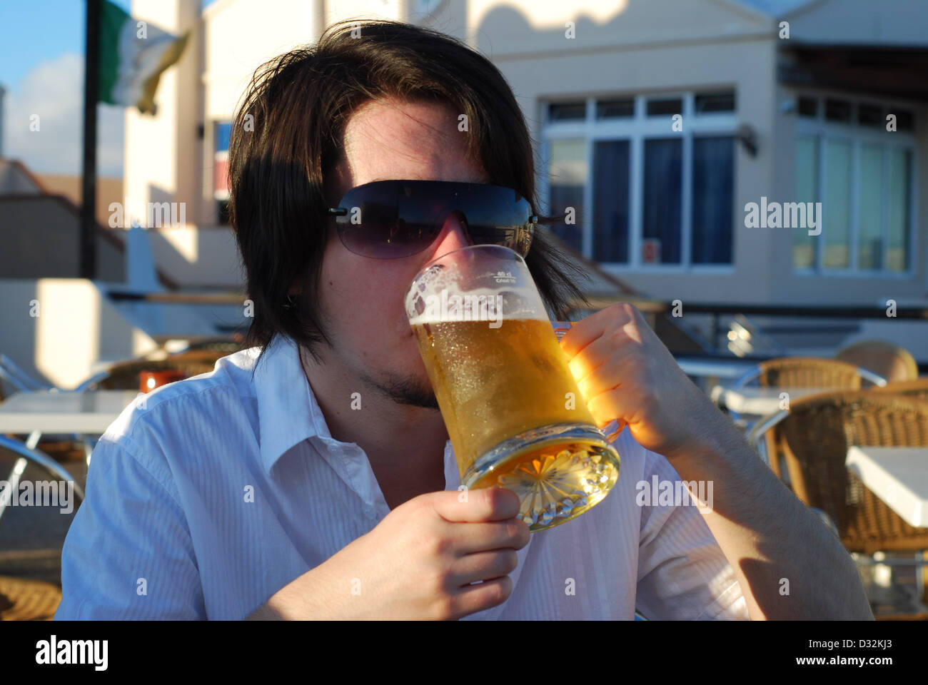 Un uomo di bere un freddo rinfrescanti bevande alcoliche su un white bar esterno tavolo in un resort per vacanze bar in calde sfumature di Sun Foto Stock