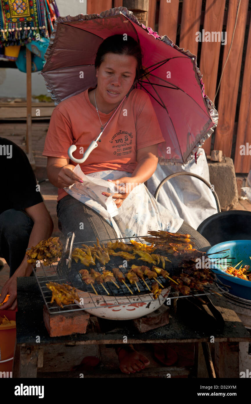 Lao donna seduta all'ombra di un ombrello al suo satay griglia cucina strada Luang prabang Laos Foto Stock