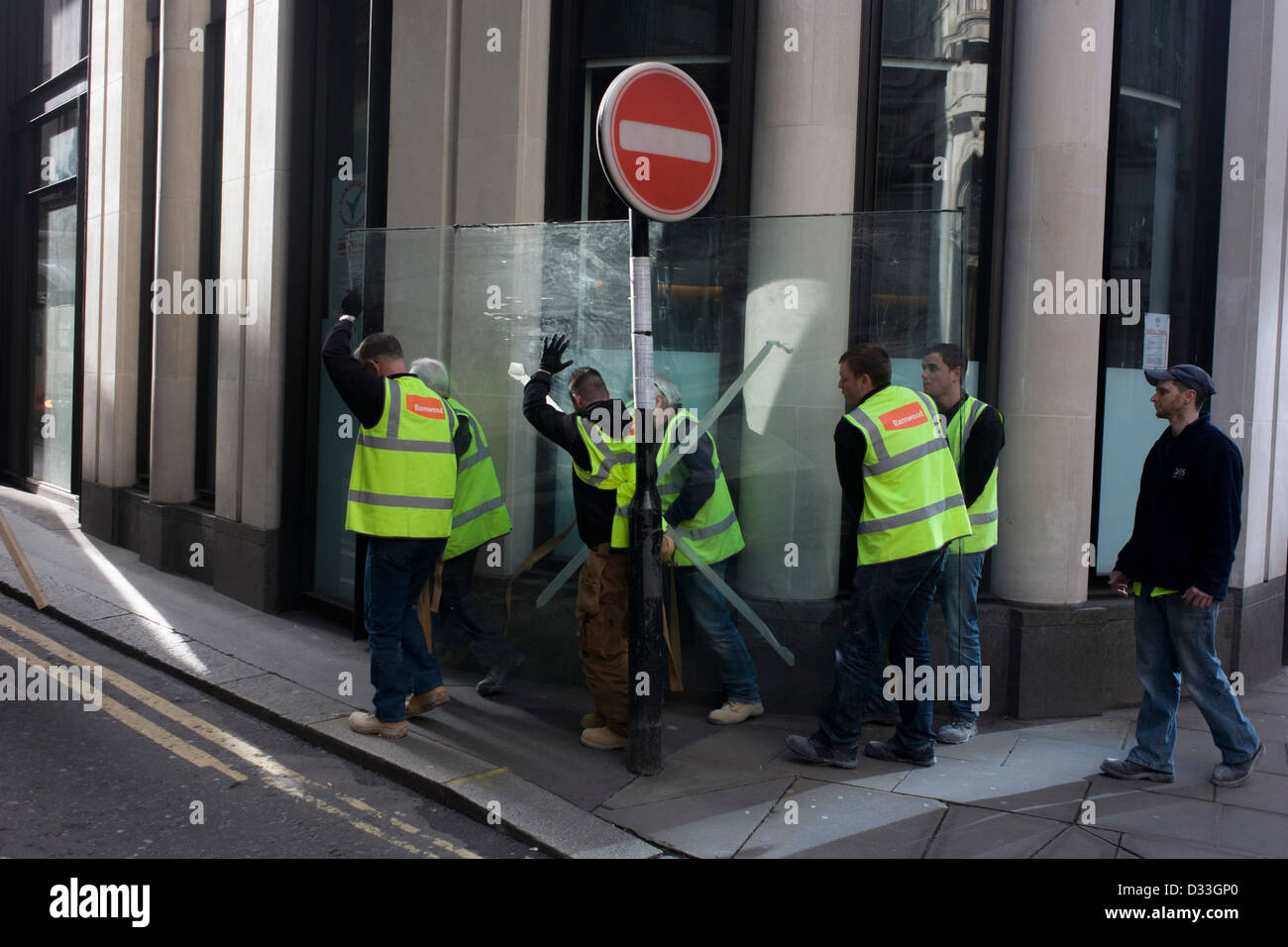 Una squadra di operai che indossa hi-visibilità tabards con il nome della società di costruzioni Barnwood sul retro, lotta per manhandle un molto pesanti a piastra di vetro attraverso una città di London street. Passando sotto una voce di alcun segno di traffico, gli uomini camminare sul marciapiede verso un camion parcheggiato che attende il suo carico successivo nel cuore della capitale del distretto finanziario altrimenti noto come Square Mile, dopo il suo volteggiare muro romano. Foto Stock