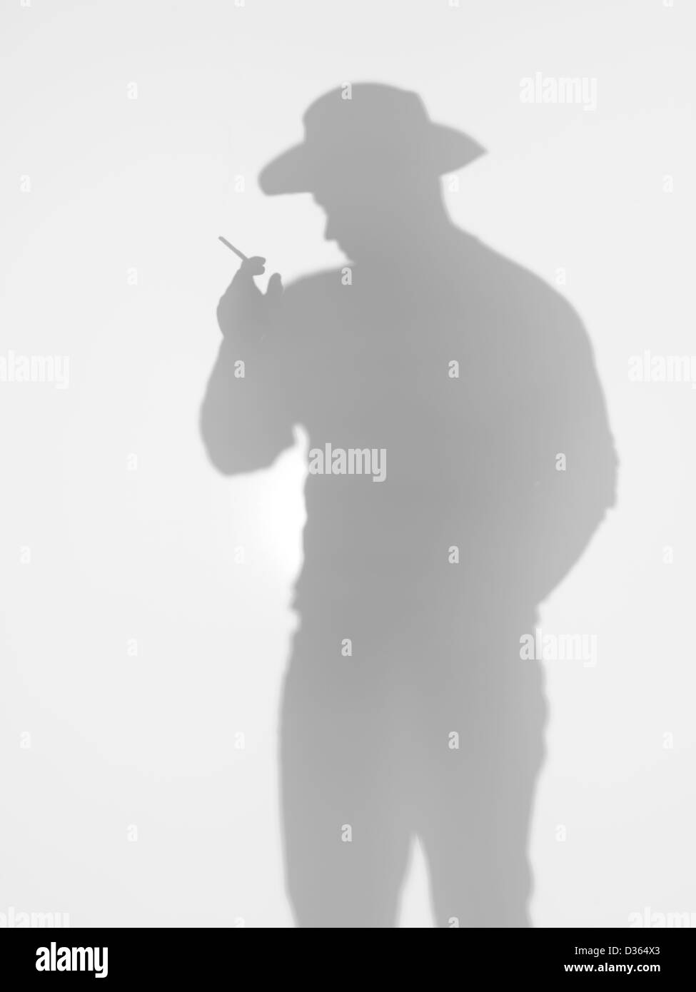 Silhouette uomo in piedi e in posa con un cappello da cowboy e fumare una sigaretta dietro una superficie diffusa Foto Stock