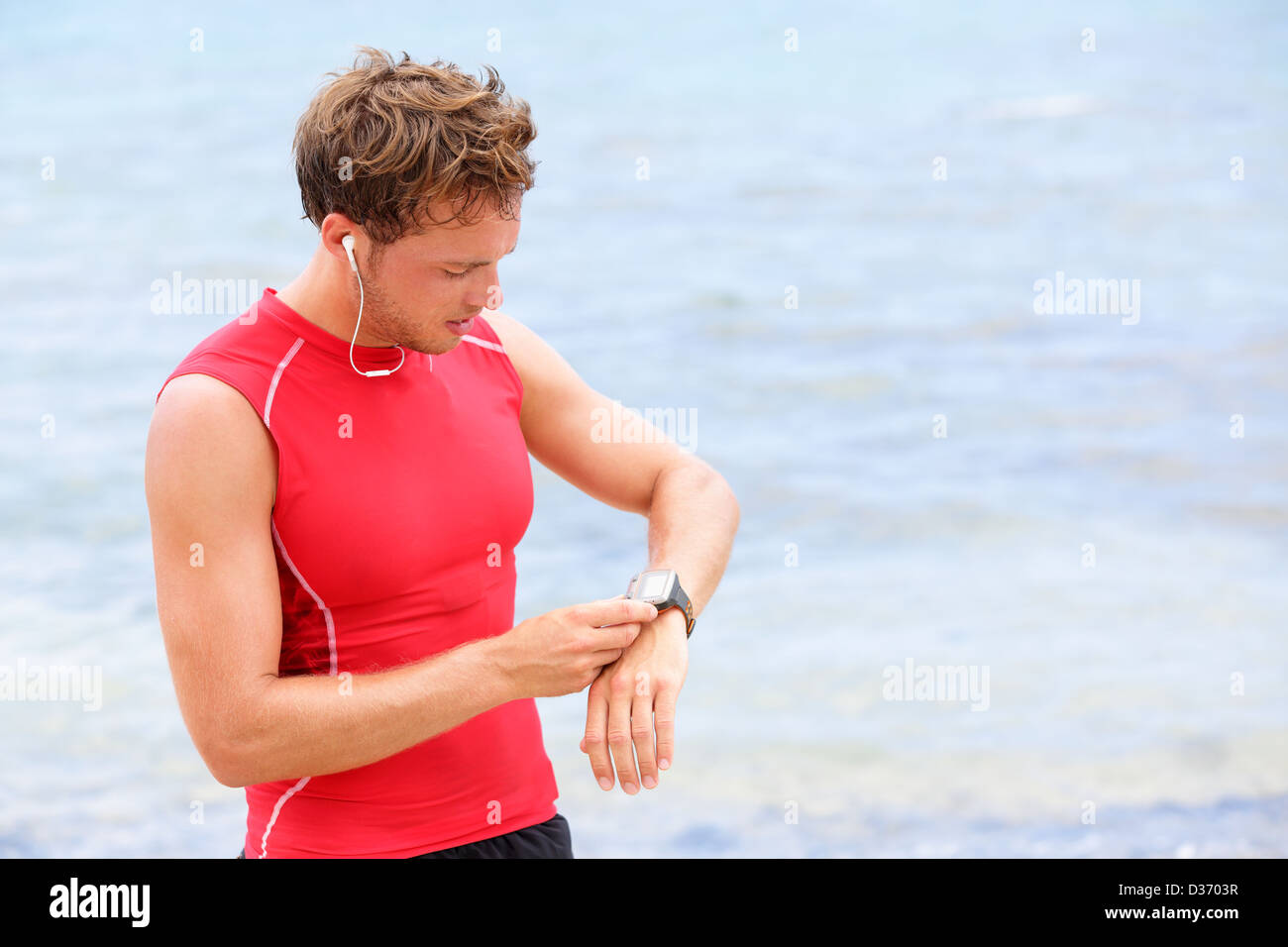 Atleta runner guardando il monitor per la frequenza cardiaca guarda. Uomo che corre sulla spiaggia prendendo una pausa in compressione t-shirt top Foto Stock