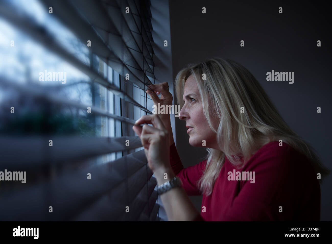 Profilo di una donna con capelli lunghi biondi, il peering attraverso una finestra cieco dall'interno guardando fuori, luce sta colpendo il suo volto. Foto Stock