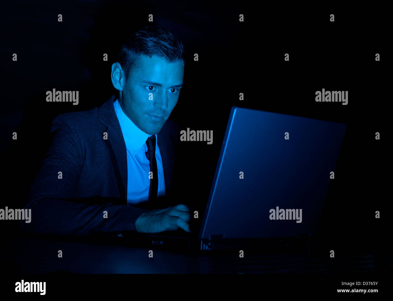 Un sospetto in cerca uomo in un business suit lavorando su un computer portatile nel buio. Potrebbe essere un hacker, o il governo snooper Foto Stock