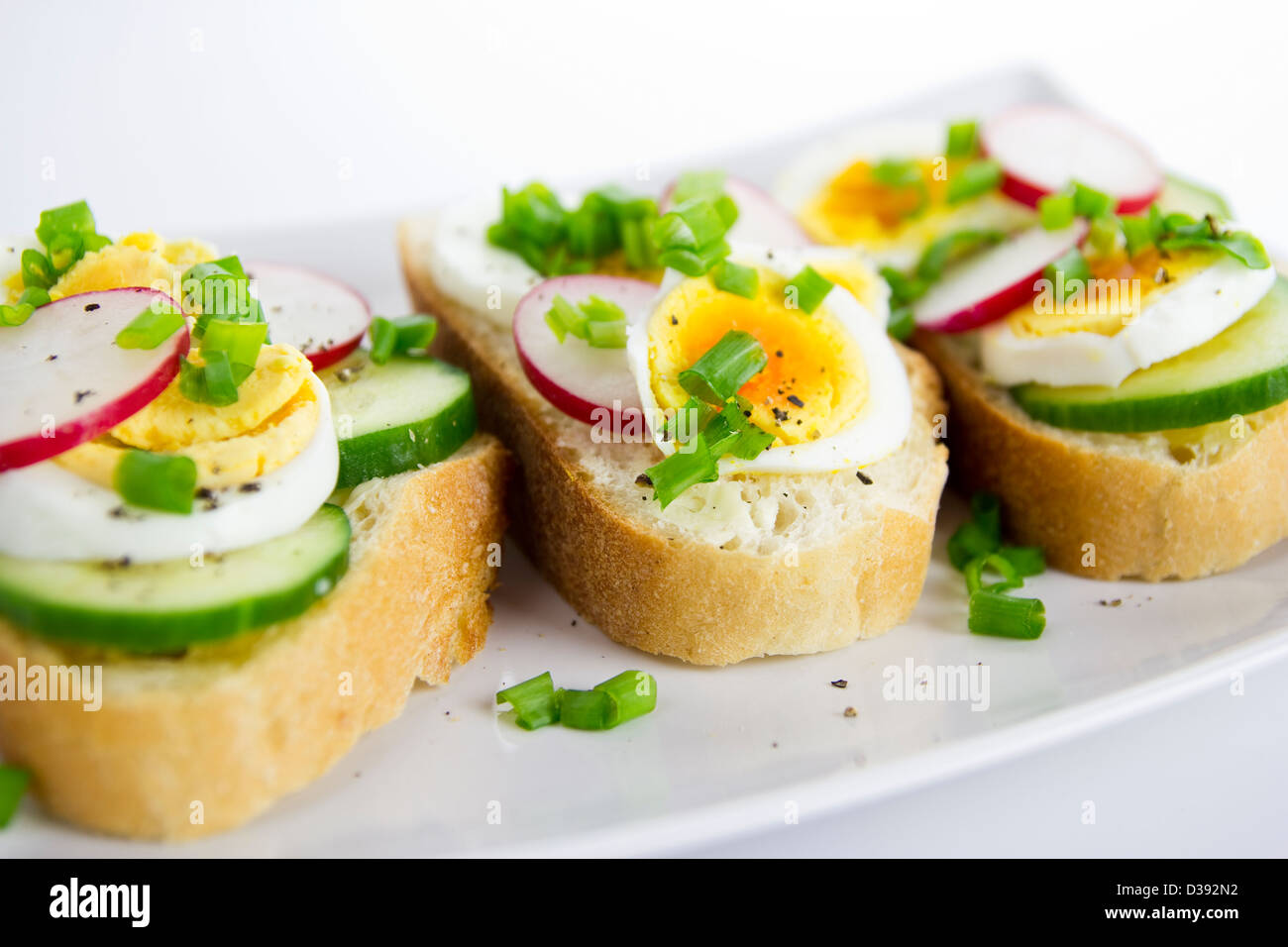 Tre panini freschi con uova, ravanello cetriolo e erba cipollina sul bianco, piastra rettangolare Foto Stock