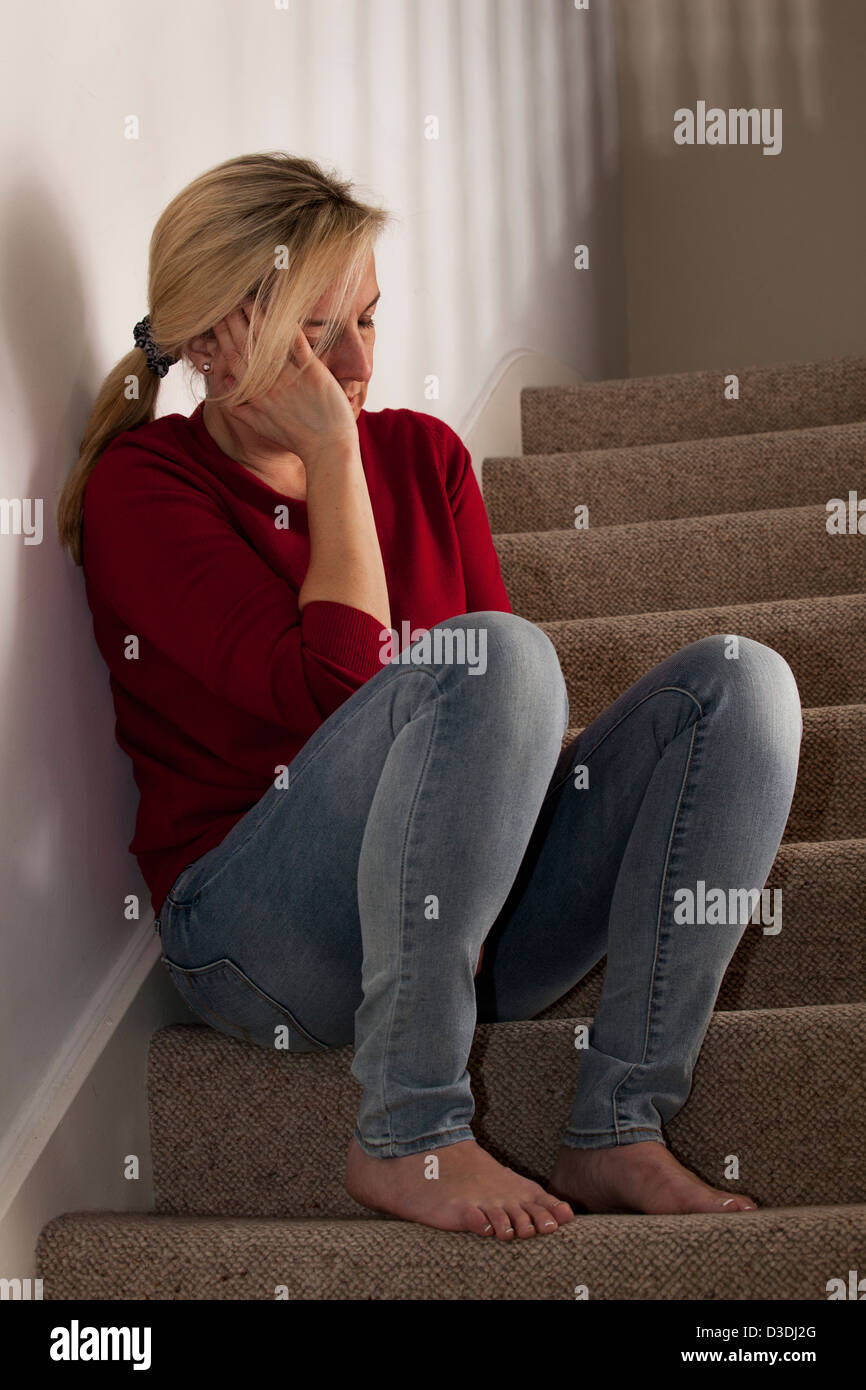 Donna bionda seduta da soli sui gradini, a casa, mano sul lato del suo viso sconvolto. Foto Stock