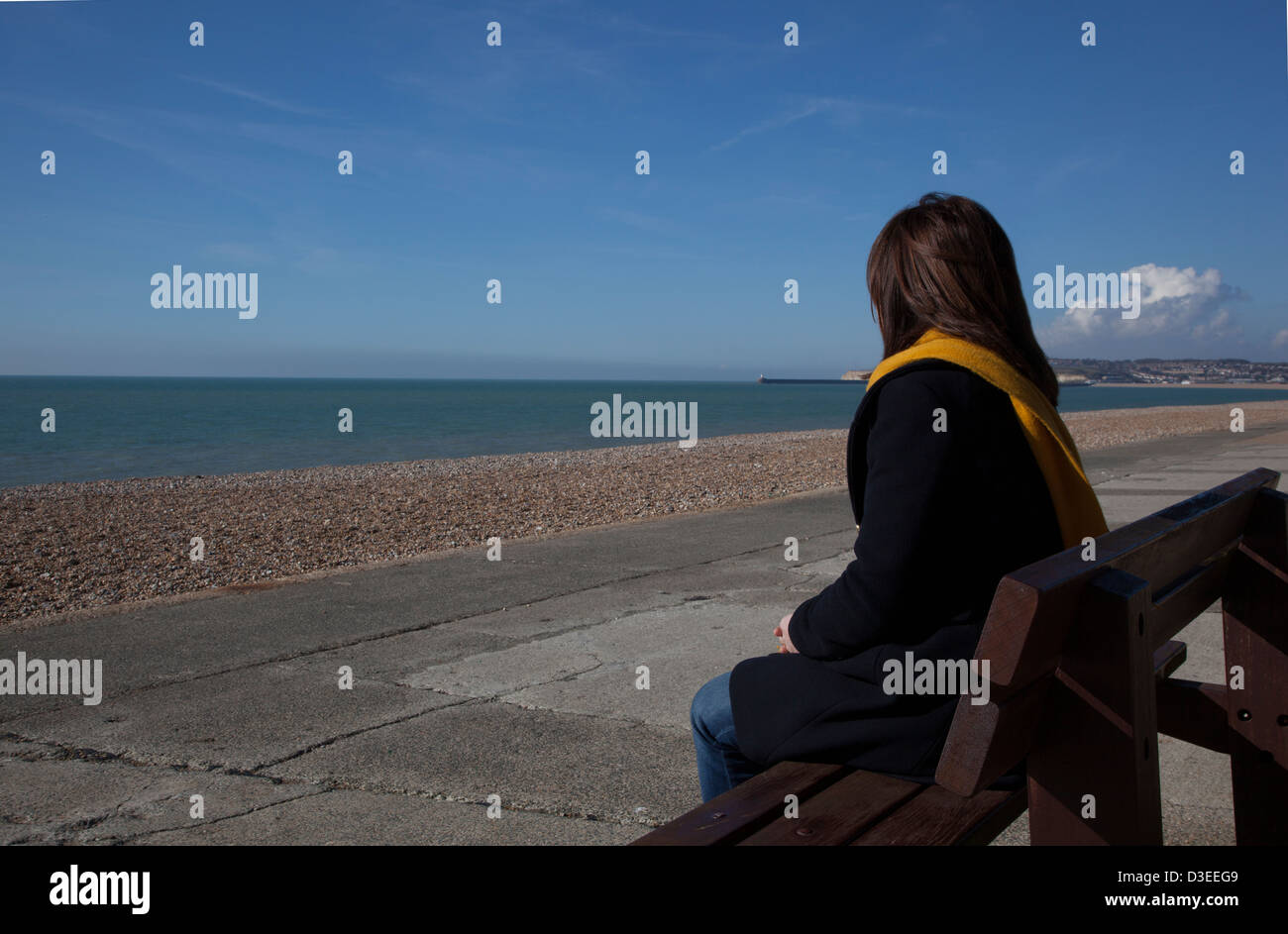 Torna alla telecamera colpo di una donna Cappotto e sciarpa seduto da solo guardando al mare, a significare la perdita di una persona cara. Foto Stock
