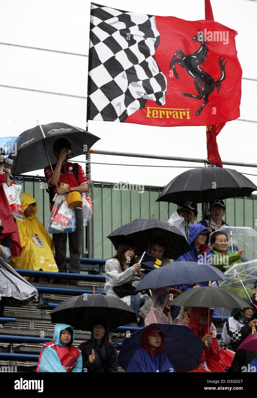 (Dpa) - Formula One fans sedersi sotto gli ombrelloni di fronte una Ferrari bandiera soffiando nel vento forte a Suzuka International Racing Course a Suzuka, in Giappone, 8 ottobre 2004. A causa di un avvicinamento typhoon, racing potrebbe essere annullato il sabato ed eventualmente anche la domenica. Il Gran Premio del Giappone si svolgerà domenica 10 ottobre 2004. Foto Stock