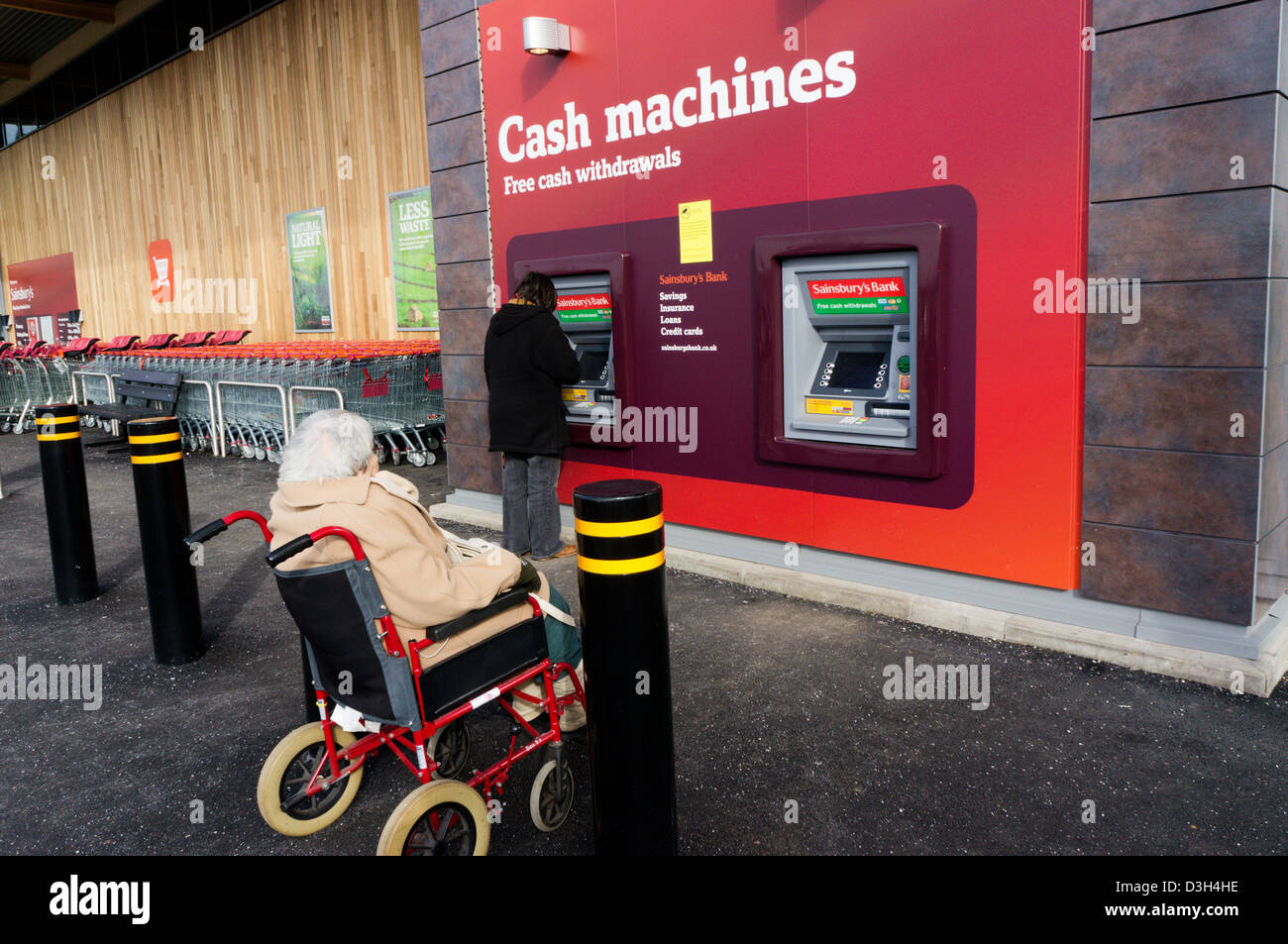 Una signora anziana è in attesa in una sedia a rotelle mentre il suo accompagnatore o assistente preleva denaro da un bancomat in un supermercato. Foto Stock