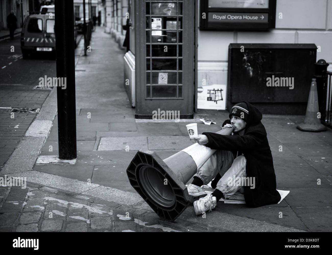 Uomo seduto sul suolo giocando un cono stradale come un corno musicale per denaro accanto al Royal Opera House, Londra Foto Stock