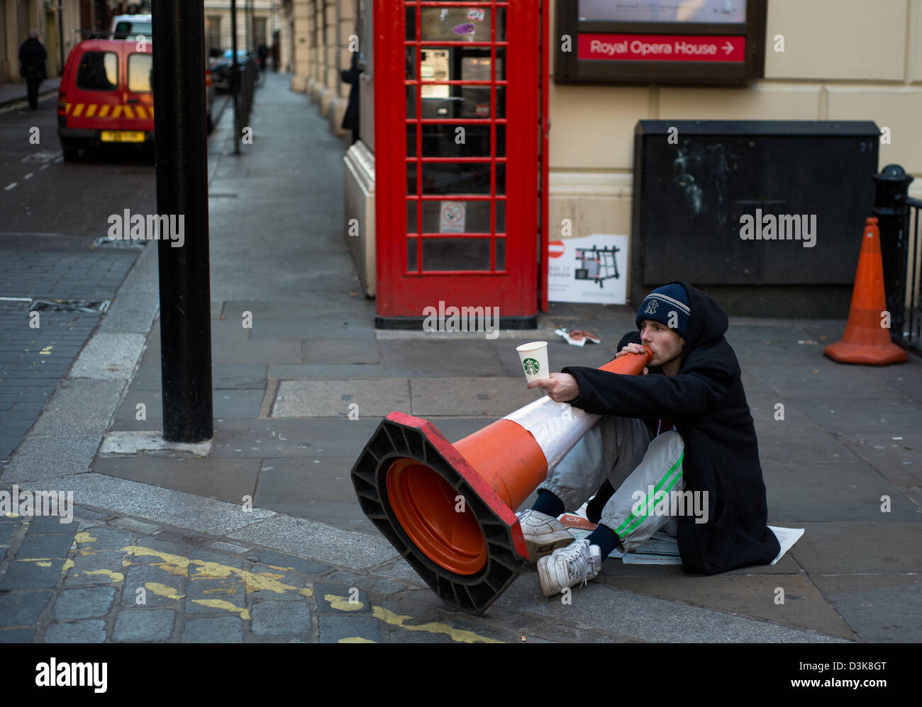 Uomo seduto sul suolo giocando un cono stradale come un corno musicale per denaro accanto al Royal Opera House, Londra Foto Stock