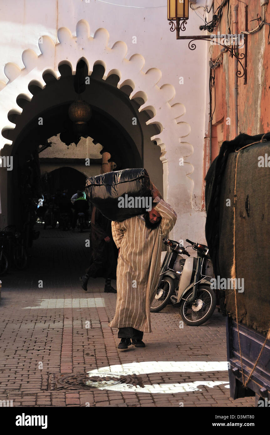 Uomo marocchino indossando un djellaba e trasportare un pesante pacchetto nero sulla sua spalla lungo una strada nella Medina di Marrakech, Marocco Foto Stock