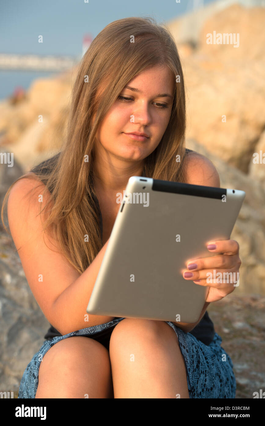 Giovane ragazza bionda seduta sulla spiaggia di rock e lavorando con i tablet pc Foto Stock