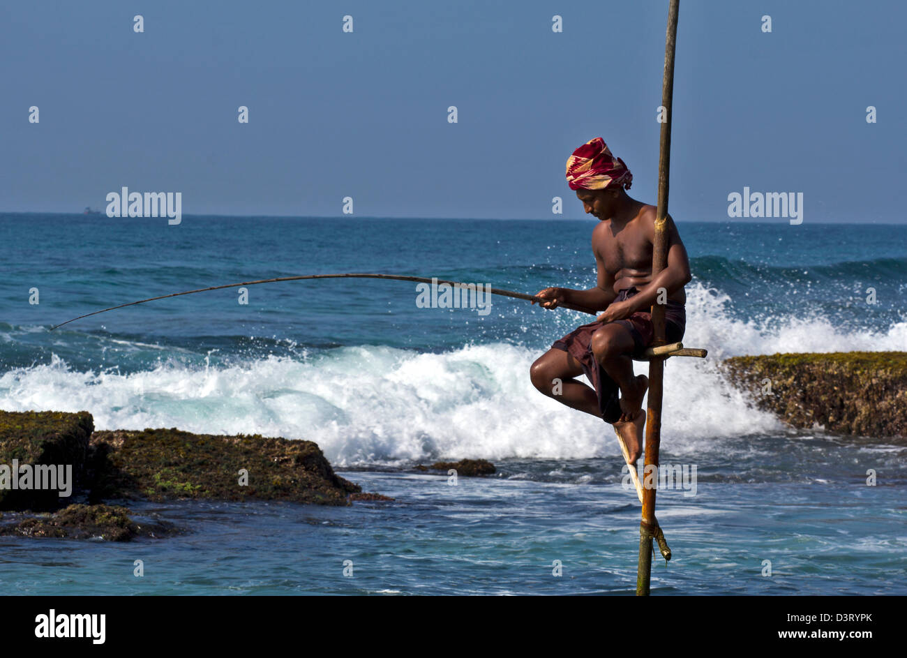 STILT PESCATORE DELLO SRI LANKA CON ONDE la battitura della roccia nelle vicinanze Foto Stock