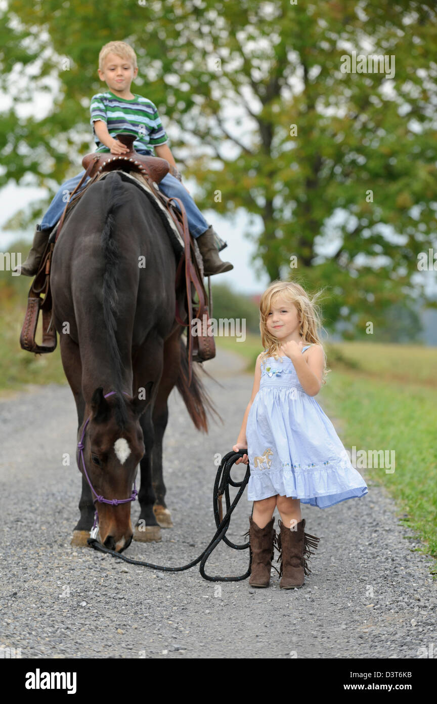 Due bambini con un grande cavallo su un vicolo del paese in estate,  fratello di equitazione e sorellina camminando in vestito blu e stivali  Foto stock - Alamy