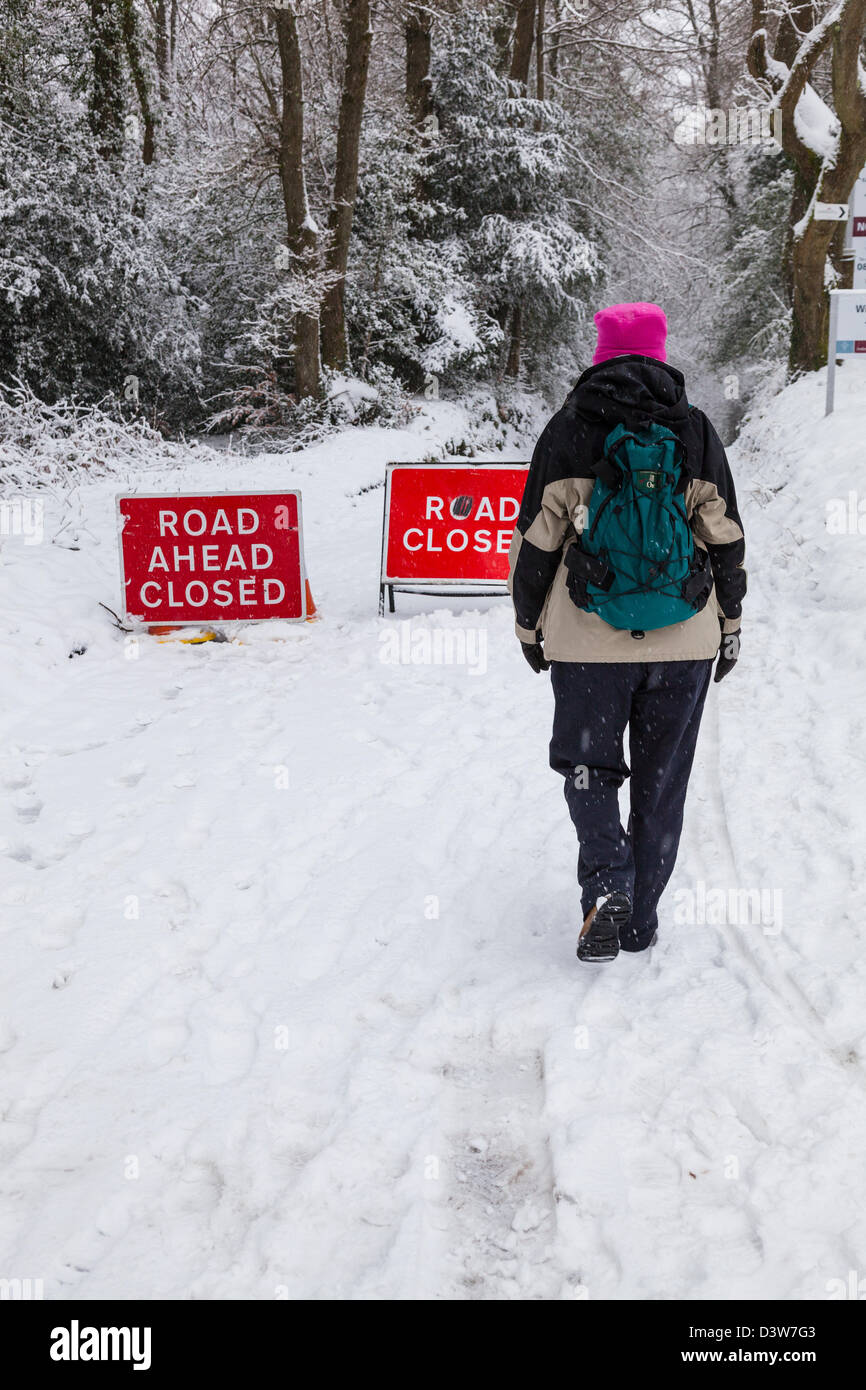 Una Donna vestita per passeggiate invernali nella neve passato "strada chiusa" segni su una coperta di neve paese LANE, Surrey, Inghilterra. Foto Stock