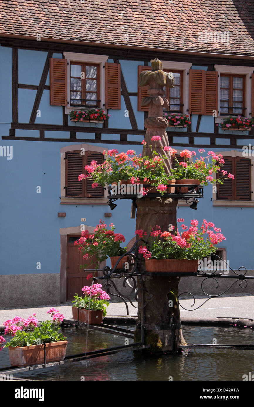 Fontana del villaggio con il blu casa in legno e muratura in Alsazia, Francia Foto Stock