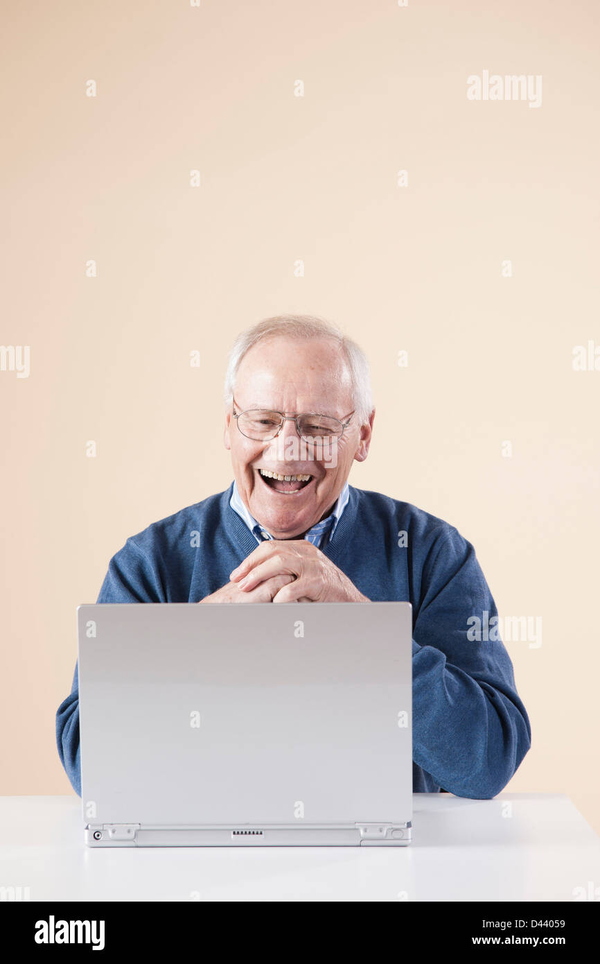 Senior uomo seduto a tavola guardando al computer portatile di ridere, Studio shot su sfondo beige Foto Stock