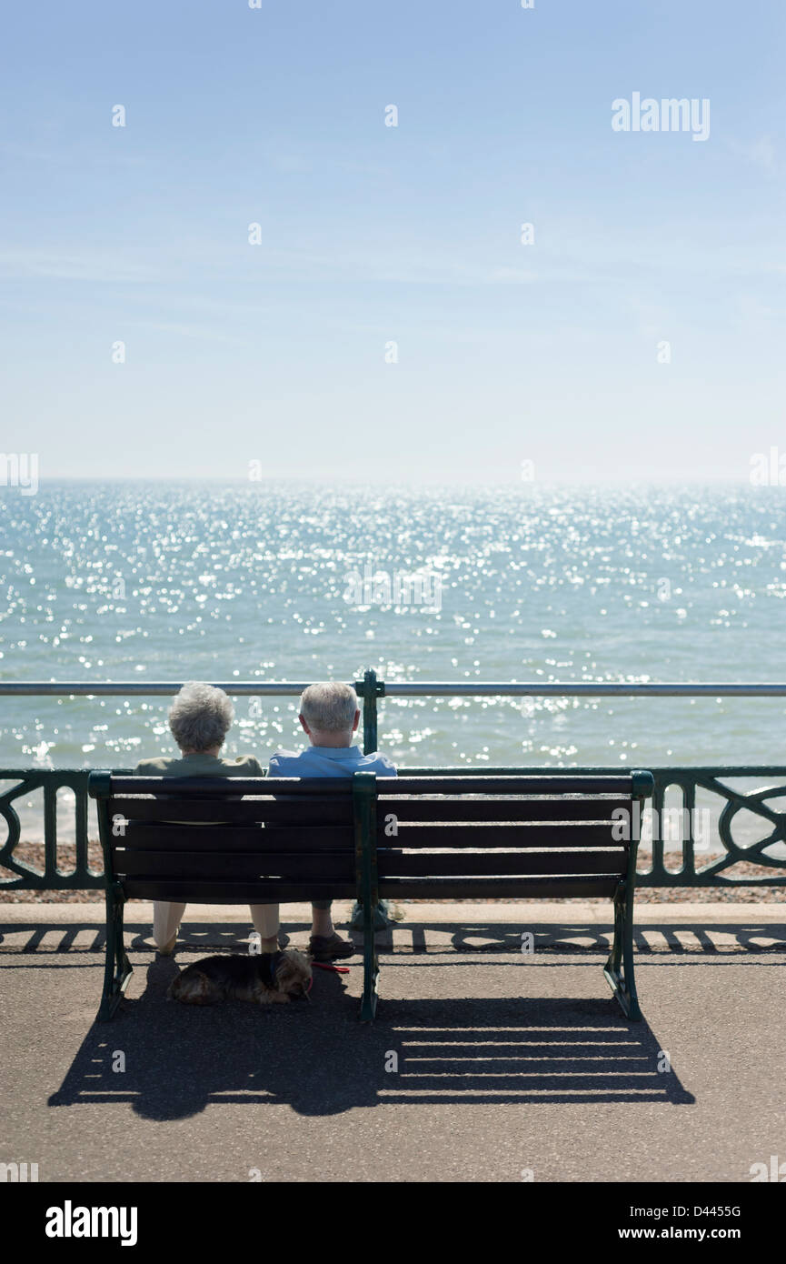Anziani vecchia coppia su un banco di lavoro per godersi il sole in riva al mare mentre il loro cane giace sotto all'ombra, REGNO UNITO Foto Stock