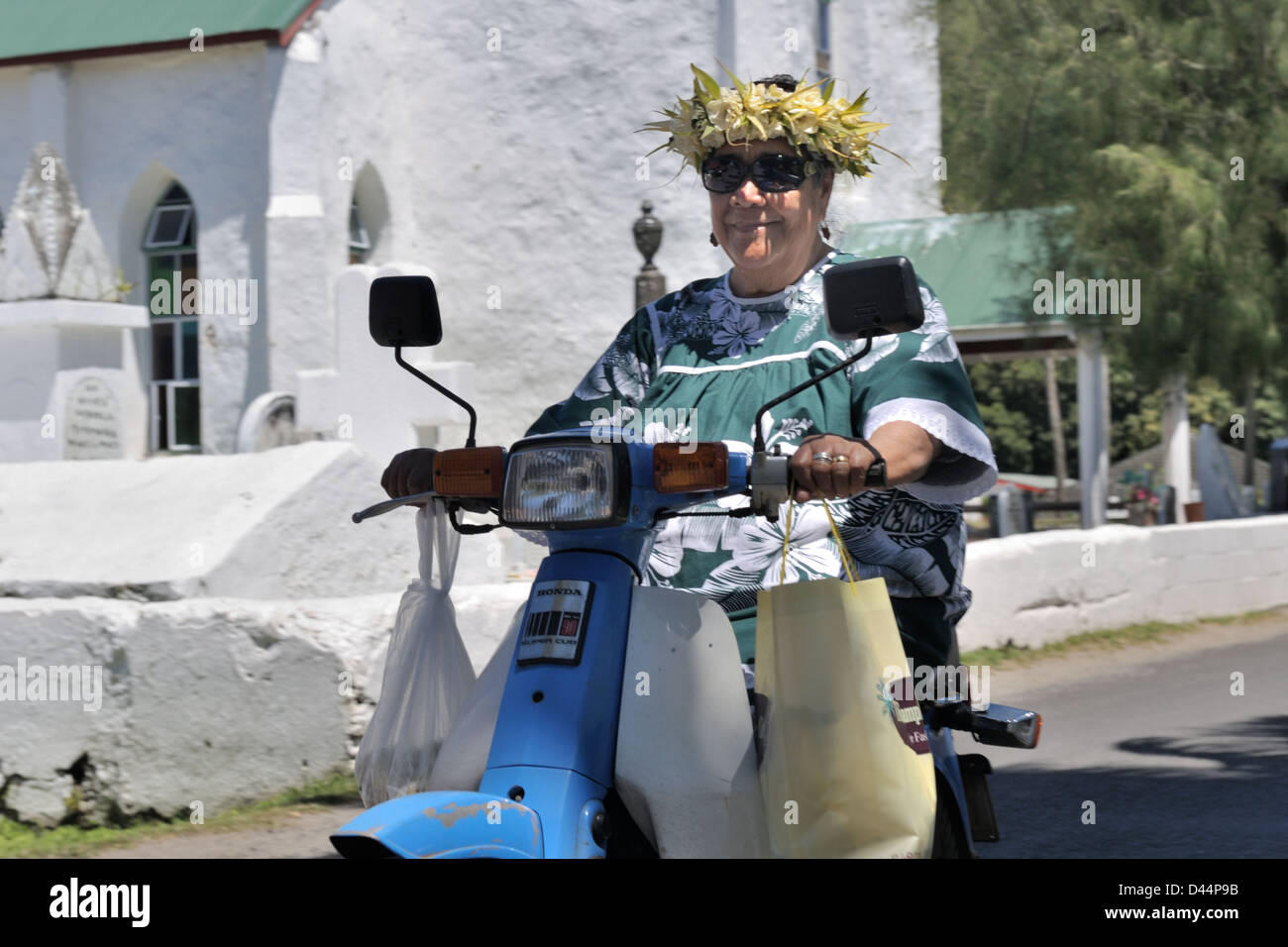 La donna in Madre Hubbard e vestito con una ghirlanda di fiori sulla sua testa, ma senza un casco a cavallo di uno scooter dopo il Vangelo giorno, Isole Cook Foto Stock