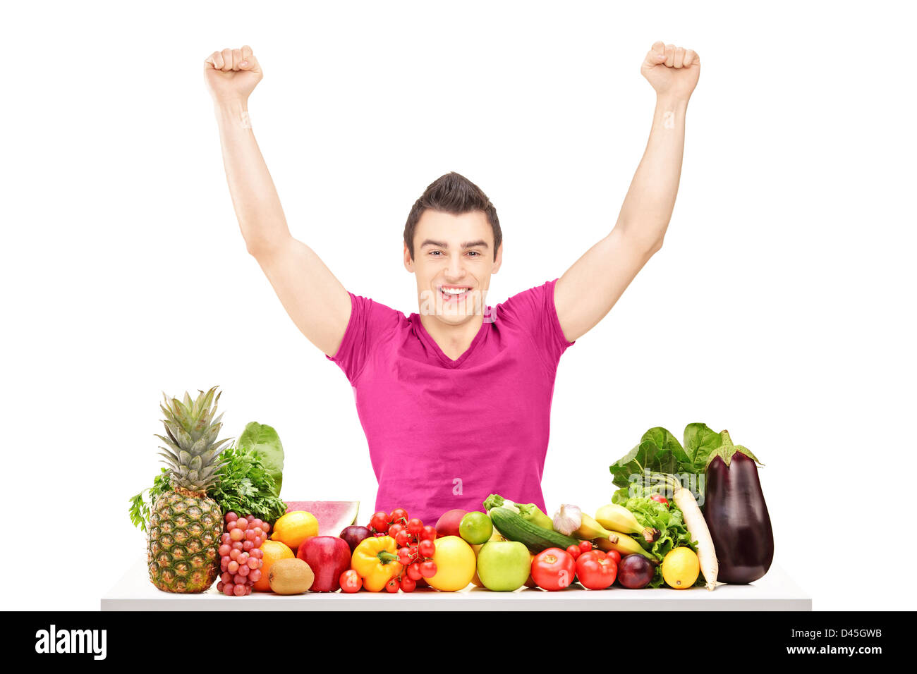 Felice giovane alzando le mani e seduto dietro un mucchio di frutta e verdura isolati su sfondo bianco Foto Stock