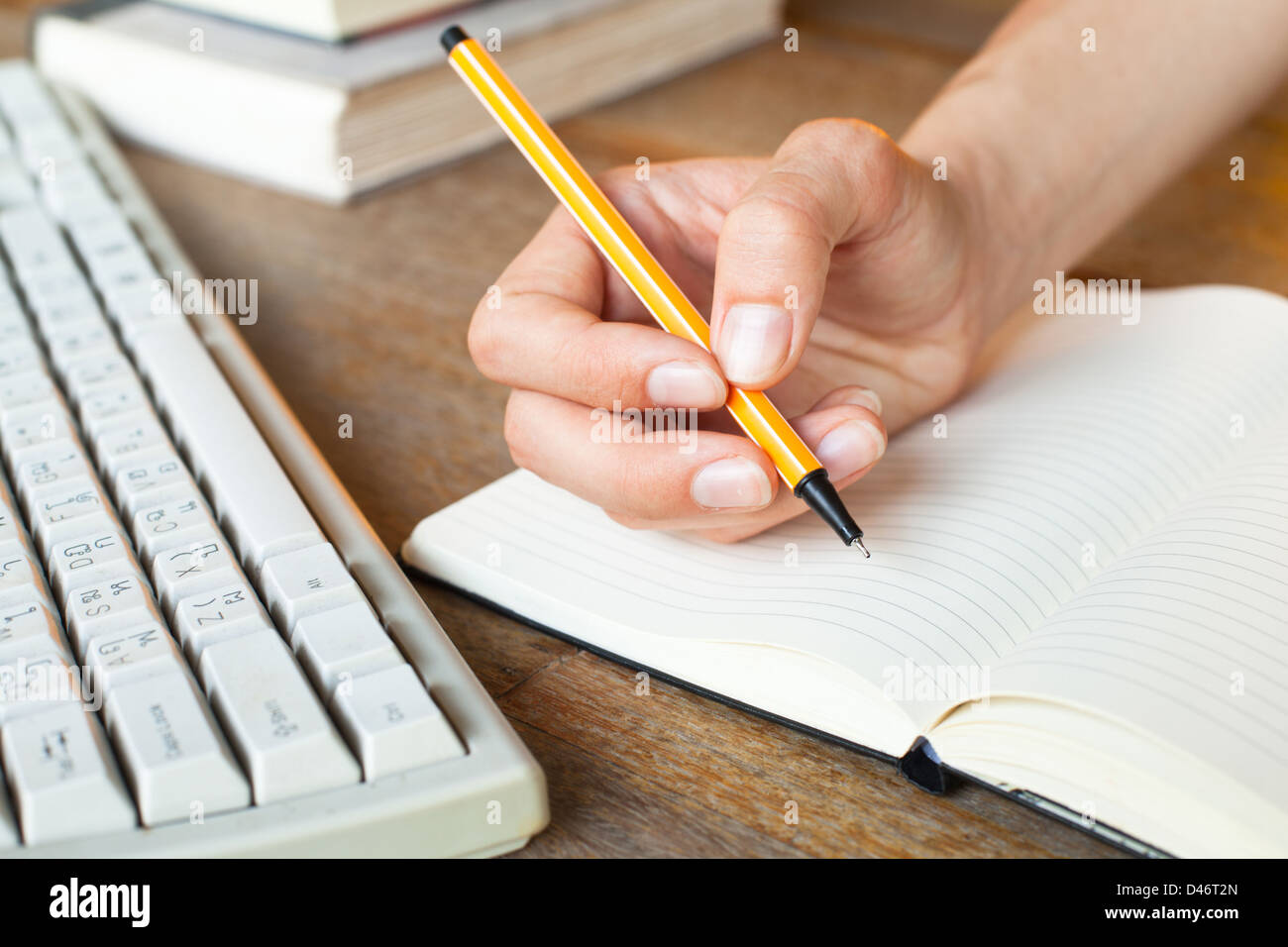 Giovane donna mani scrive una penna in un notebook, la tastiera del computer e una pila di libri in background Foto Stock