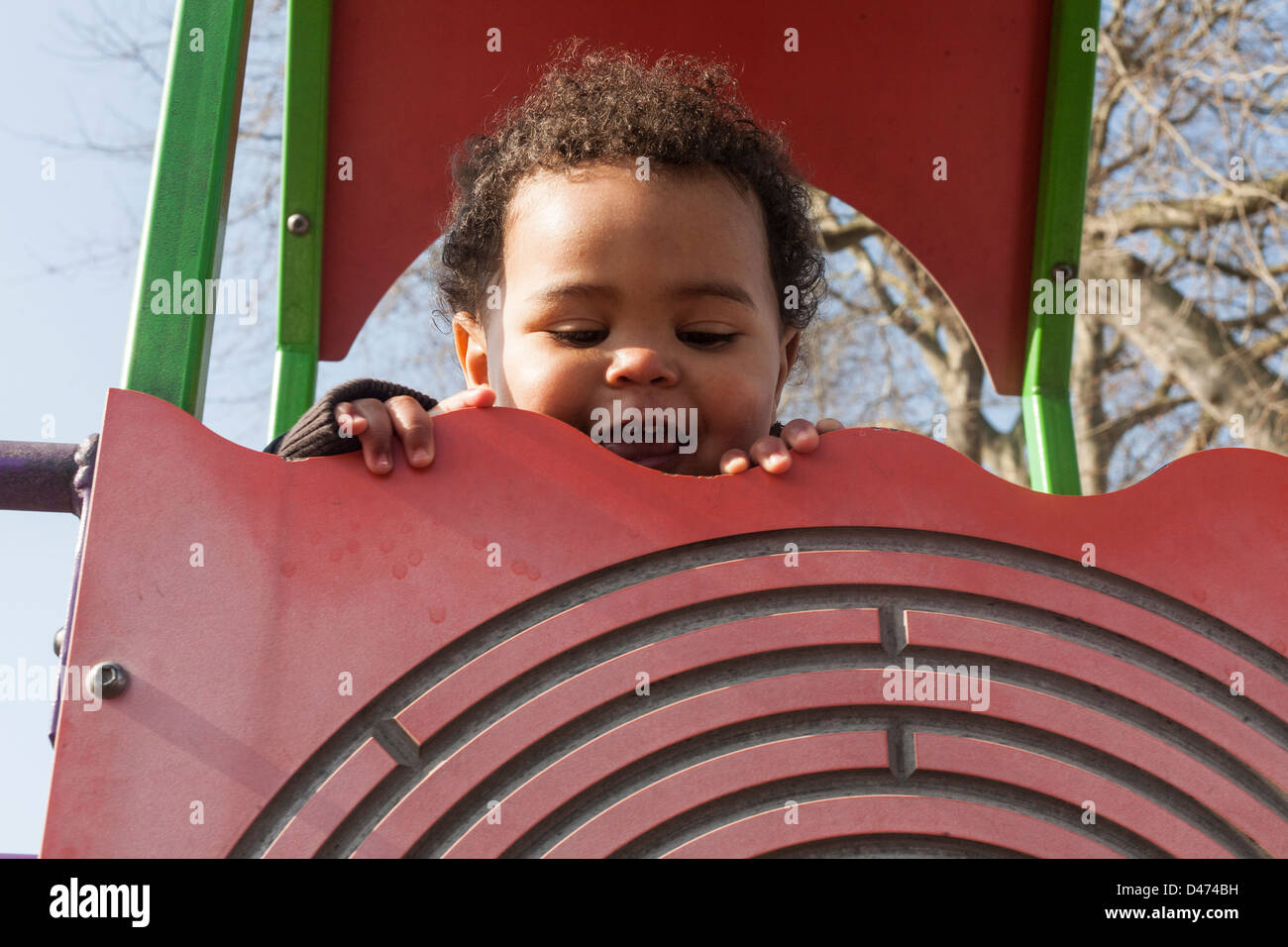 Razza mista toddler baby sorridente e giocando sul telaio di arrampicata in una giornata di sole peeking sopra la parte superiore della parete Foto Stock