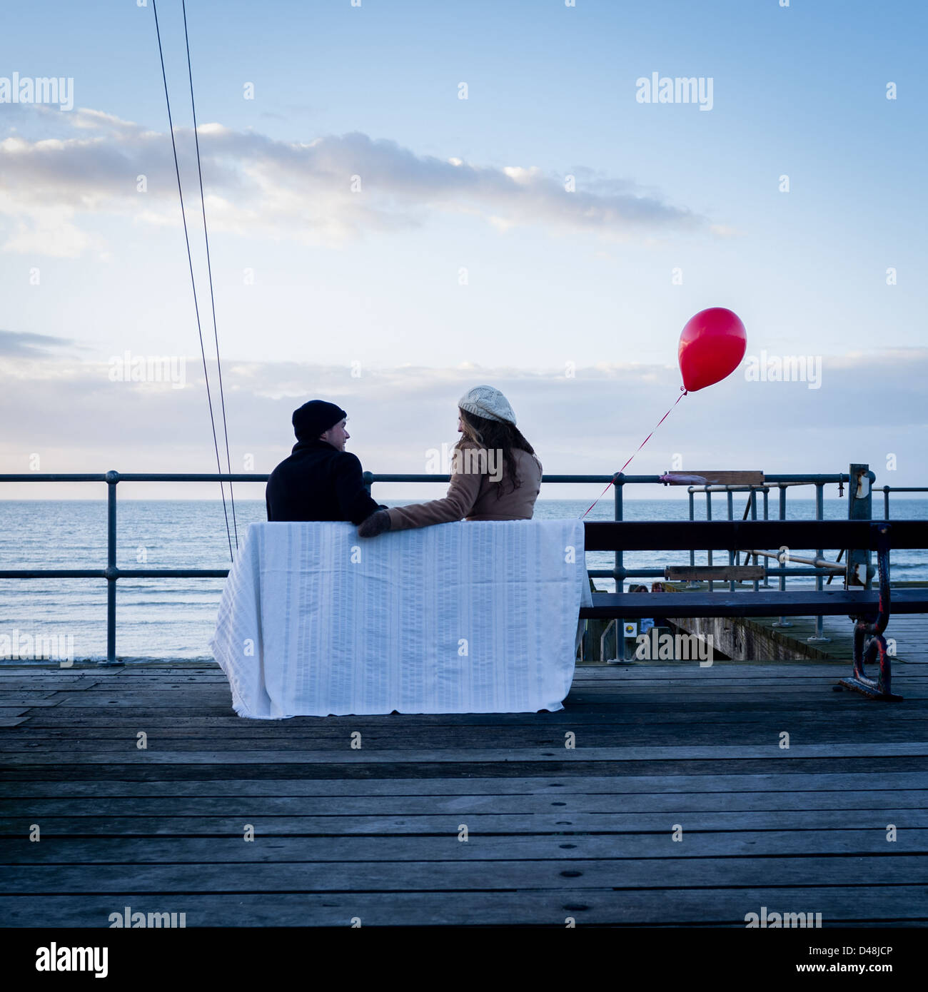 Vista posteriore di una giovane coppia seduta su una panchina sul mare con un palloncino rosso il giorno di San Valentino Feb 14 REGNO UNITO Foto Stock