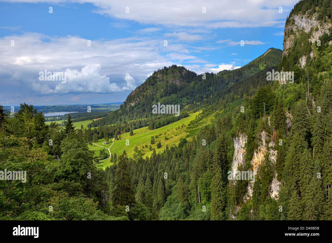 Vista del soleggiato verde prato circondato da monti coperti di boschi sotto il bellissimo cielo molto nuvoloso in Baviera, Germania. Foto Stock