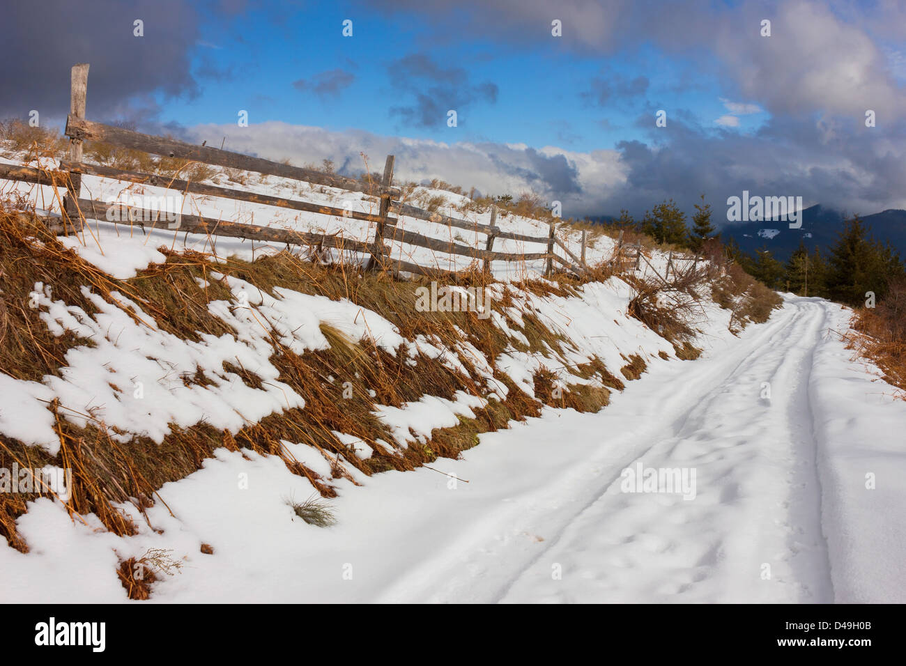 Colpo in prospettiva della rurale snowy stretta strada di alta montagna, con staccionata in legno e drammatici sky in background. Foto Stock