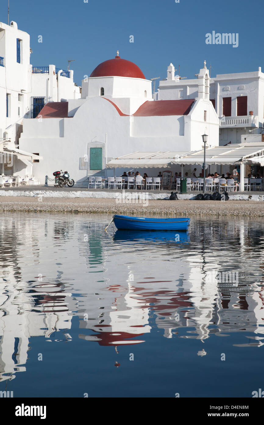 Chiesa bianca con cupola rossa, caffè all'aperto, barca e riflessi nelle acque del porto di Mykonos, isole greche Foto Stock