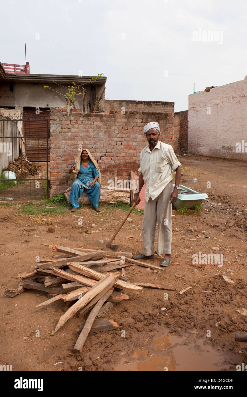 Punjabi uomo trinciatura di legno al di fuori del tradizionale casa di famiglia con sua moglie guardando Foto Stock