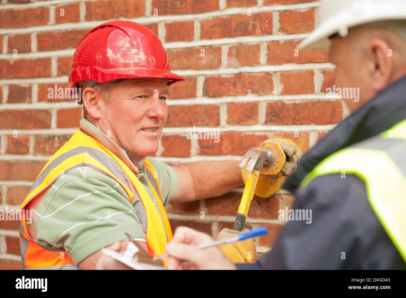 Builder facendo lavori di riparazione ad un muro di mattoni con la costruzione di ispettore cercando su e prendere appunti. Foto Stock