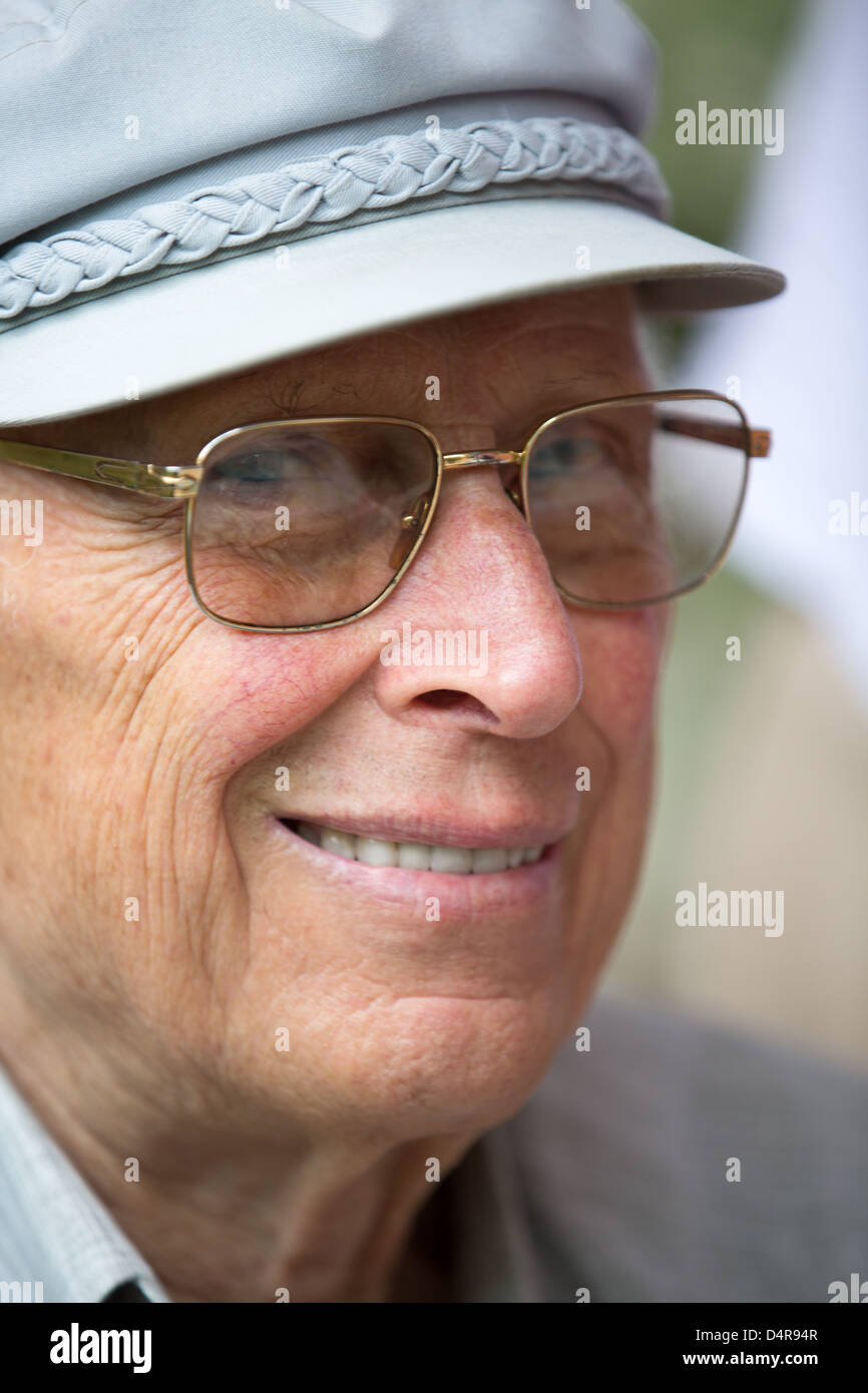 Senior è uomo sorridente in telecamera con il suo classico cappellino grigio e di colore dorato bicchieri. Foto Stock