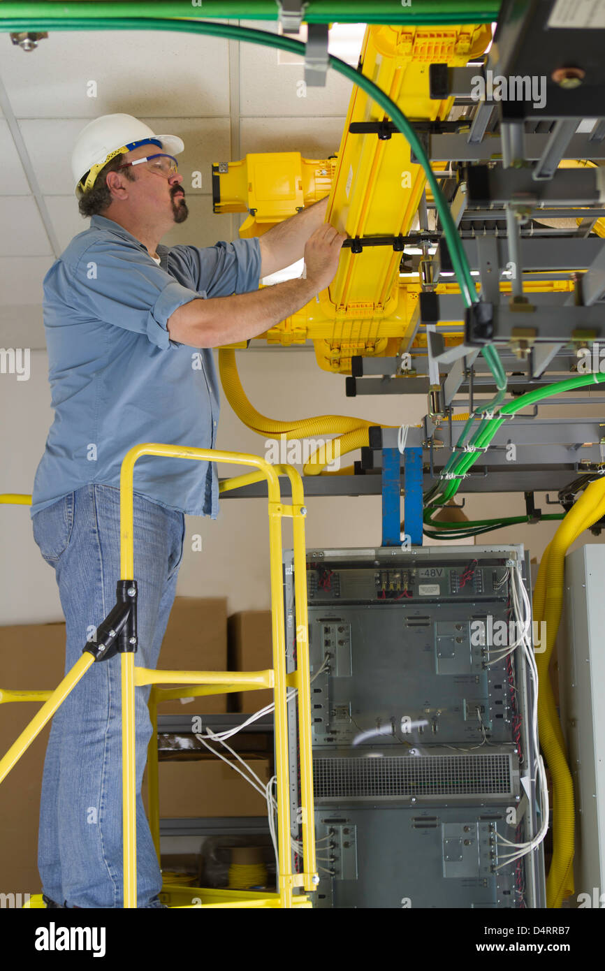 Un hardhat commutatore di telecomunicazione lavoratore di ispezionare i cavi a fibre ottiche in fibra condotto su un omologato OSHA scaletta della piattaforma. Foto Stock