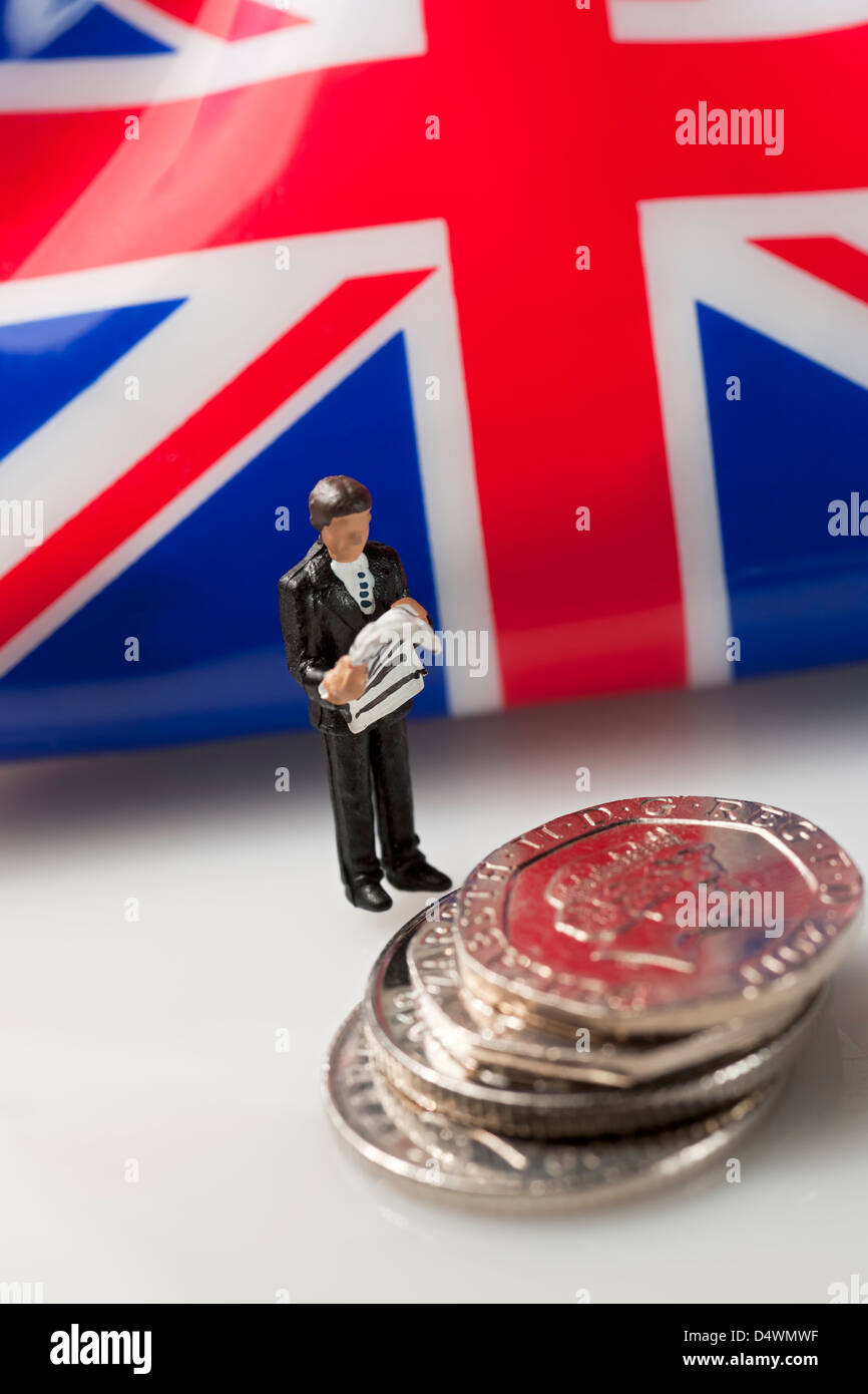 Primo piano di un giocattolo una piccola figura di uomo d'affari maleman si trovava accanto a una pila di monete inglesi da 20 pence per finanziare il concetto di denaro aziendale Foto Stock