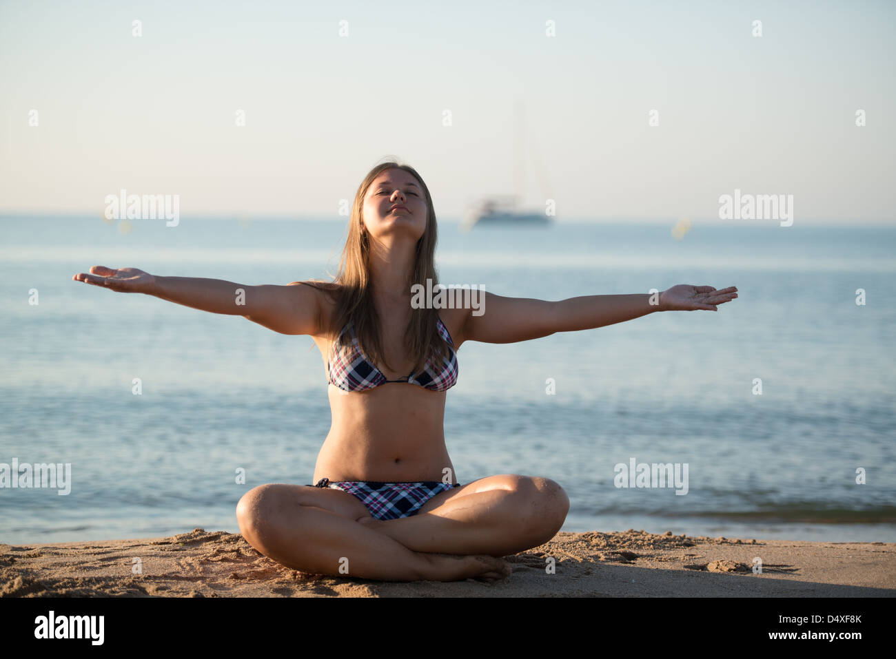 Giovane donna bionda rilassante fare yoga sulla spiaggia di sabbia di mare e yacht sullo sfondo Foto Stock