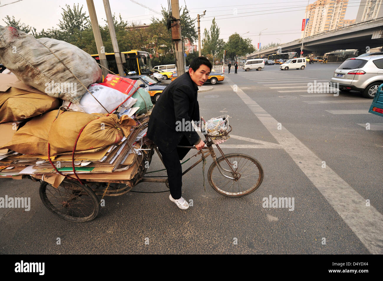 Cina, Pechino, deliveryman il trasporto del carico sulla bicicletta mentre le auto in movimento su strada con edifici in background Foto Stock