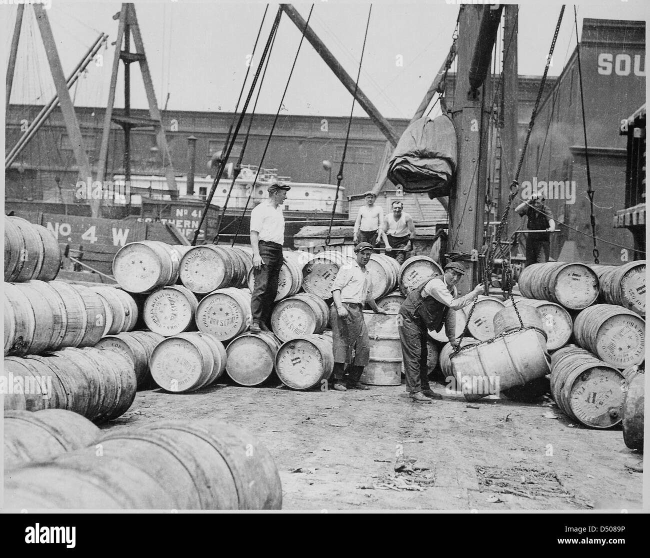 La fotografia di Lewis Hine del 1912 cattura i stevedores che caricano barili di corn Dyrup su una chiatta presso un molo di New York. Questa immagine documenta le condizioni di lavoro dei lavoratori, evidenziando le pratiche di lavoro industriale dei primi anni del XX secolo e le questioni del lavoro minorile. Foto Stock