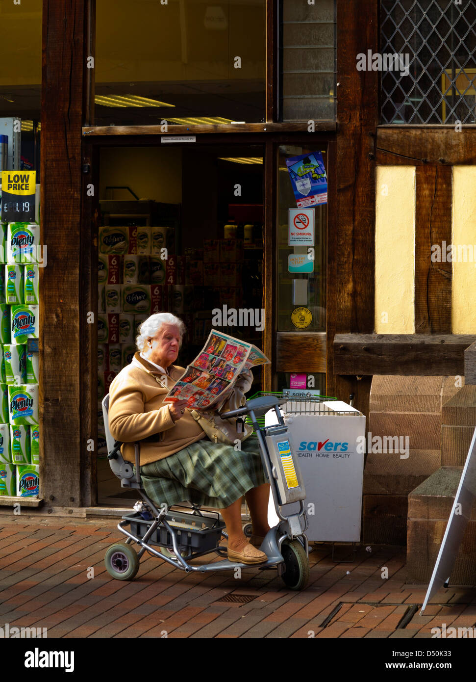 Donna anziana in scooter di mobilità leggendo un giornale al di fuori di un negozio in Stafford England Regno Unito Foto Stock
