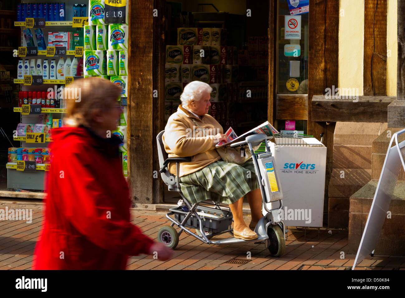 Donna anziana in scooter di mobilità leggendo un giornale al di fuori di un negozio in Stafford England Regno Unito Foto Stock