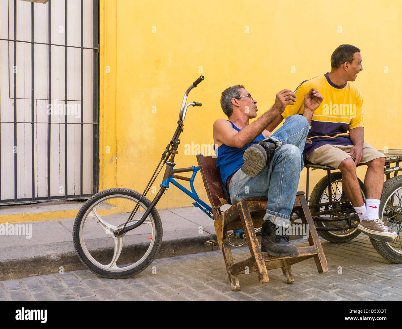 Due uomini cubani di sedersi di fronte un giallo brillante parete mediante le loro biciclette gesticolando e avente una riscaldata conversazione in Havana, Cuba Foto Stock