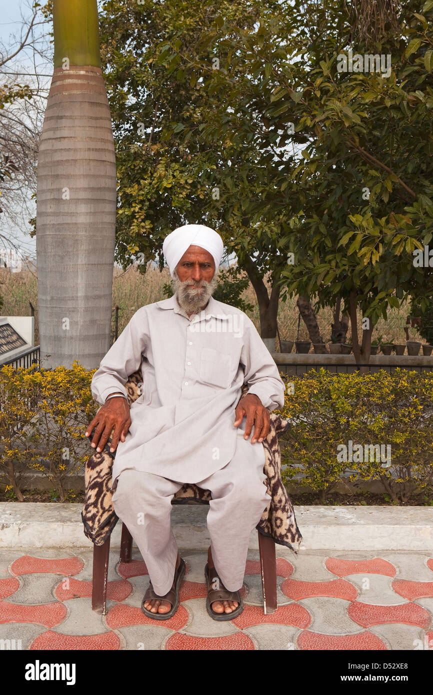 Un anziano Sikh seduto in una sedia da un albero di palma su un cortile pavimentato in Gurdwara a Harike in Punjab (India). Foto Stock