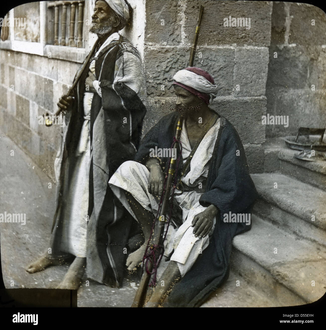 Questa fotografia cattura un gruppo di beduini arabi in abito tradizionale ad Alessandria, in Egitto. Gli uomini sono seduti e in piedi vicino a una scala, con lanterne sulla scena, offrendo uno sguardo sullo stile di vita tradizionale dei beduini nell'Egitto del XX secolo. Foto Stock