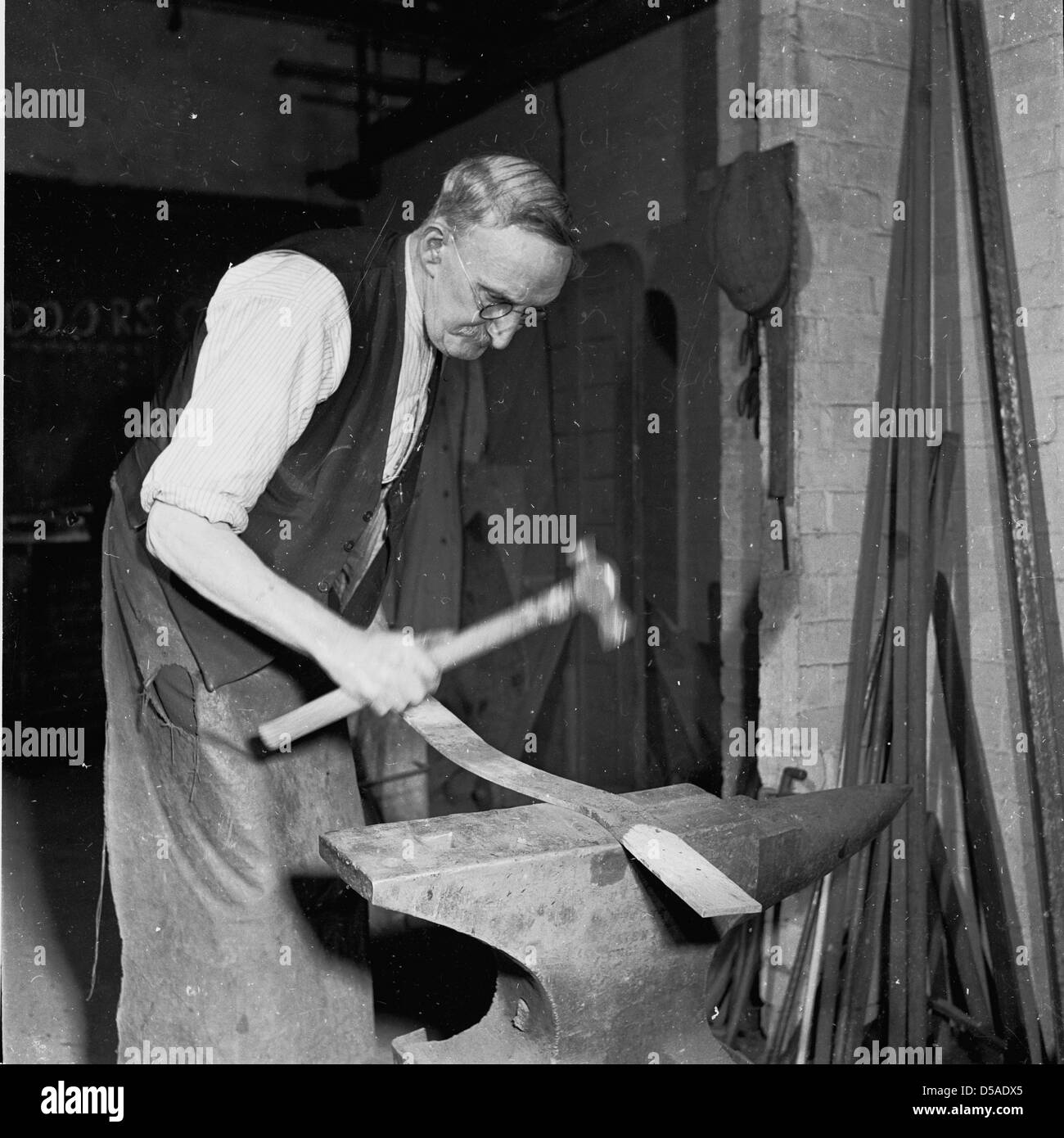 Storico, anni '50. Fabbro anziano che indossa un grembiule lungo e in un gilet usando un'incudine ed un martello per modellare un pezzo lungo di metallo. Foto Stock