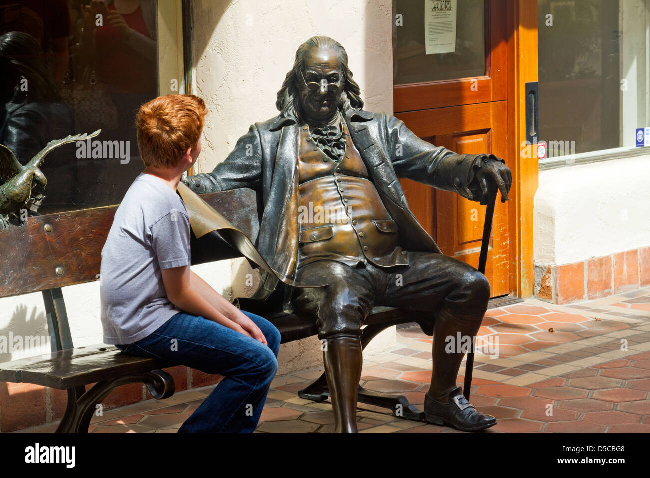 Redheaded ragazzo si siede su un banco di lavoro con la statua in bronzo di Ben Franklin a Santa Barbara in California Foto Stock