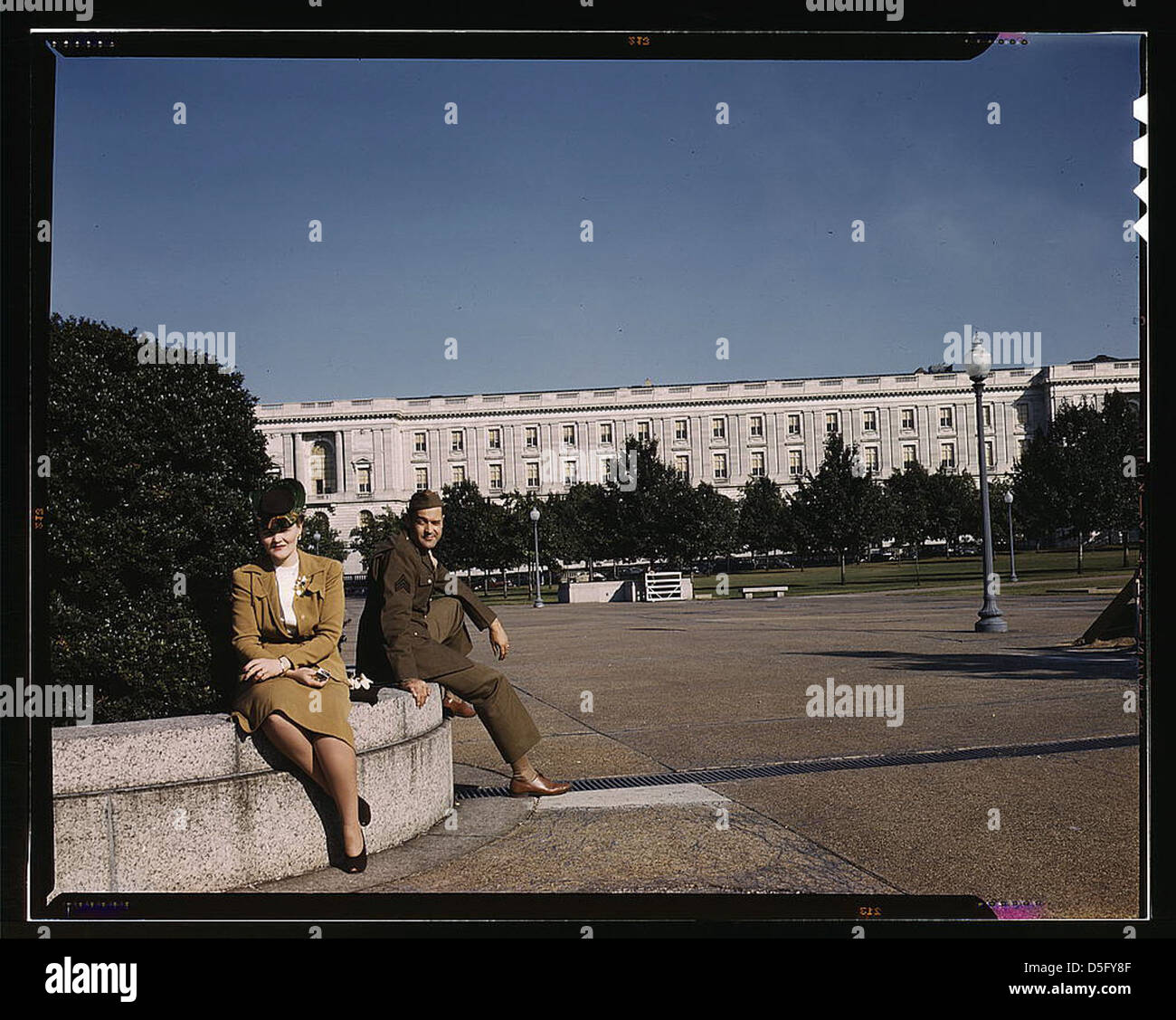 Una fotografia degli anni '1940 che mostra un soldato e una donna seduti su una panchina di un parco a Washington, D.C., con il Russell Senate Office Building sullo sfondo. L'immagine cattura l'epoca della guerra e l'architettura storica di Washington, D.C. Foto Stock