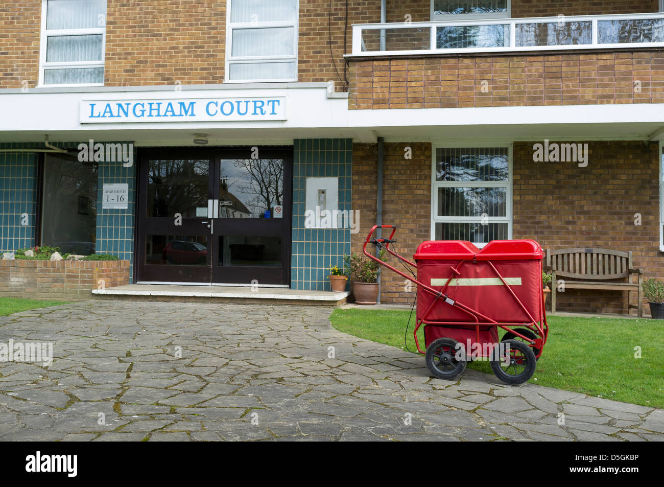 Un postino britannico il carrello al di fuori di un blocco di appartamenti mentre il postino recapita la posta all'interno. Foto Stock