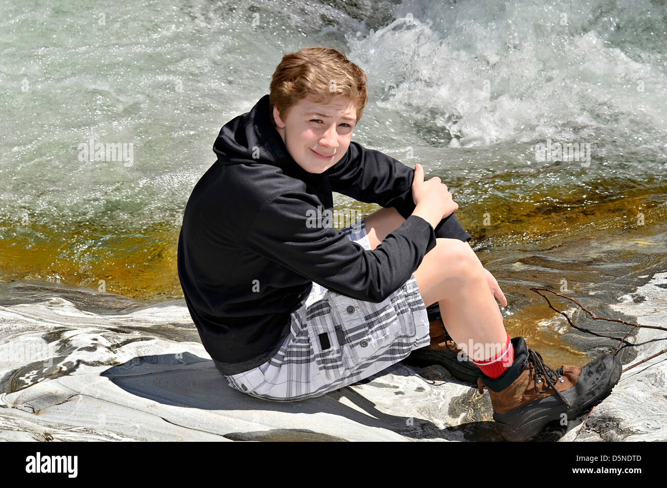 Un giovane ragazzo seduto sulle rocce al lato di un fiume. Foto Stock